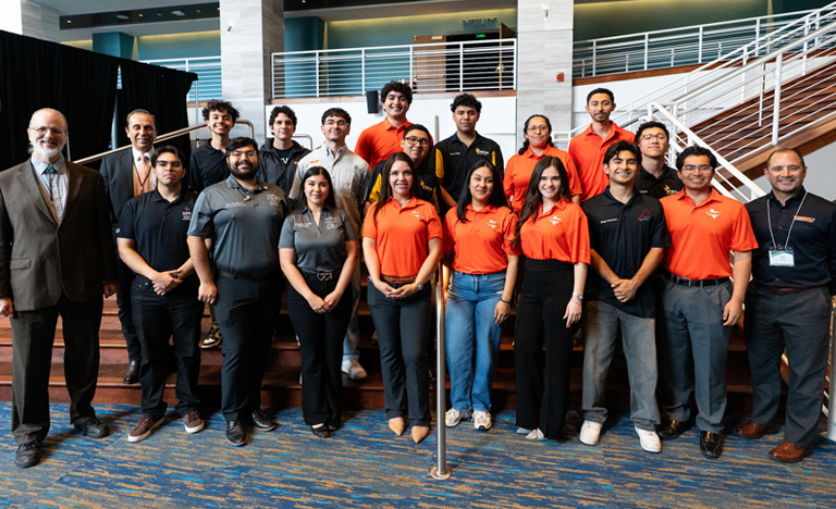 Large group of attendees posing for a photo inside a bright, multi-level indoor venue, with staircases, railings, and stage lighting visible in the background.