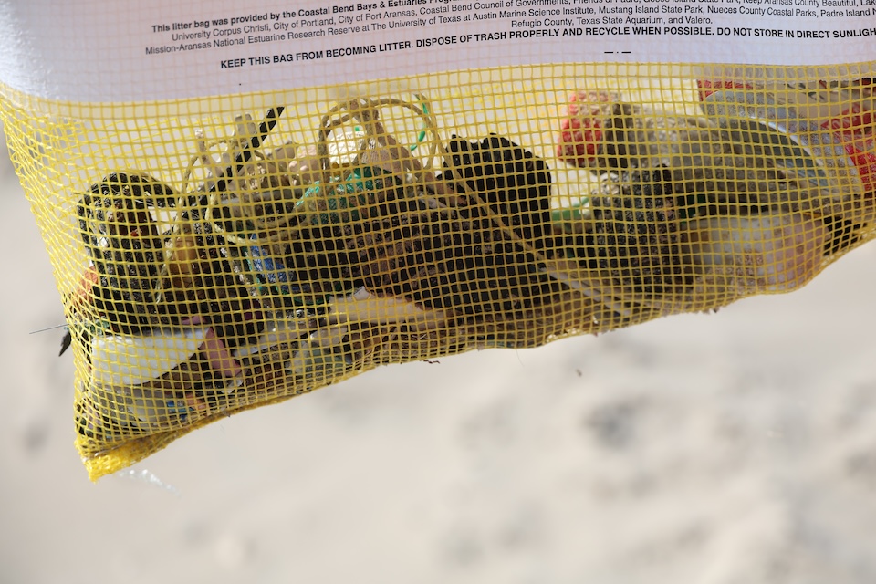 A yellow mesh trash bag filled with collected beach debris, including plastic bottles and small pieces of waste, hangs against a blurred sandy background.