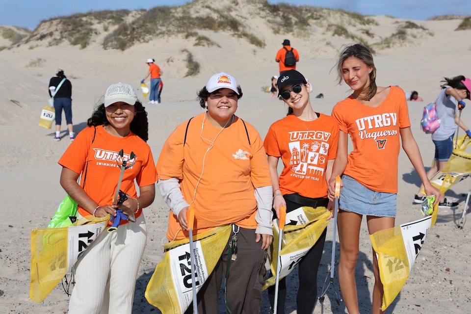 A group of UTRGV volunteers wearing orange shirts stand on the beach holding trash grabbers and yellow mesh bags during a beach cleanup event.