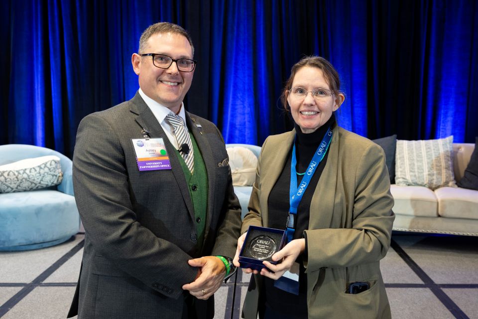 “Two attendees standing indoors on a stage, with one attendee holding a small glass award plaque