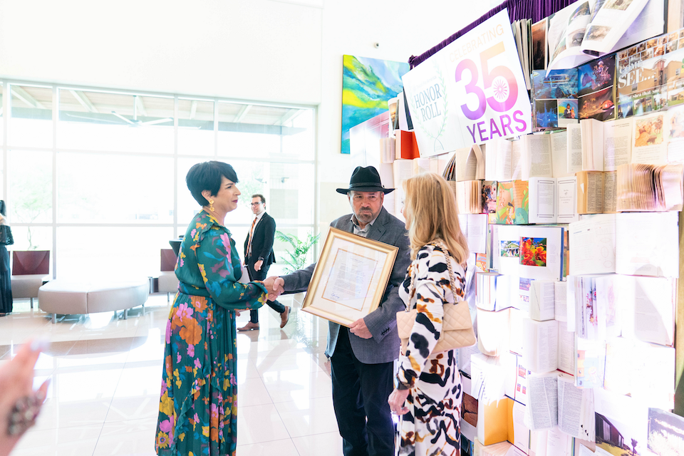 Two people exchange a framed document while standing near a wall display of pages and artwork, with a sign reading ‘35 Years’ visible above the display.