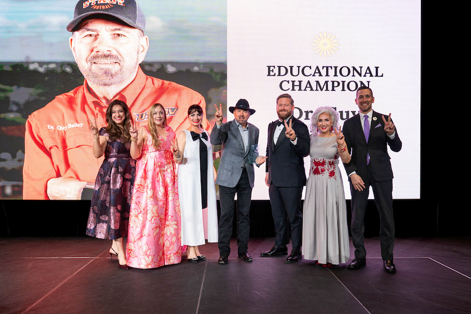 A group of attendees stand on a stage posing for a photo, with a large screen behind them showing a portrait and the words ‘Educational Champion’ partially visible.