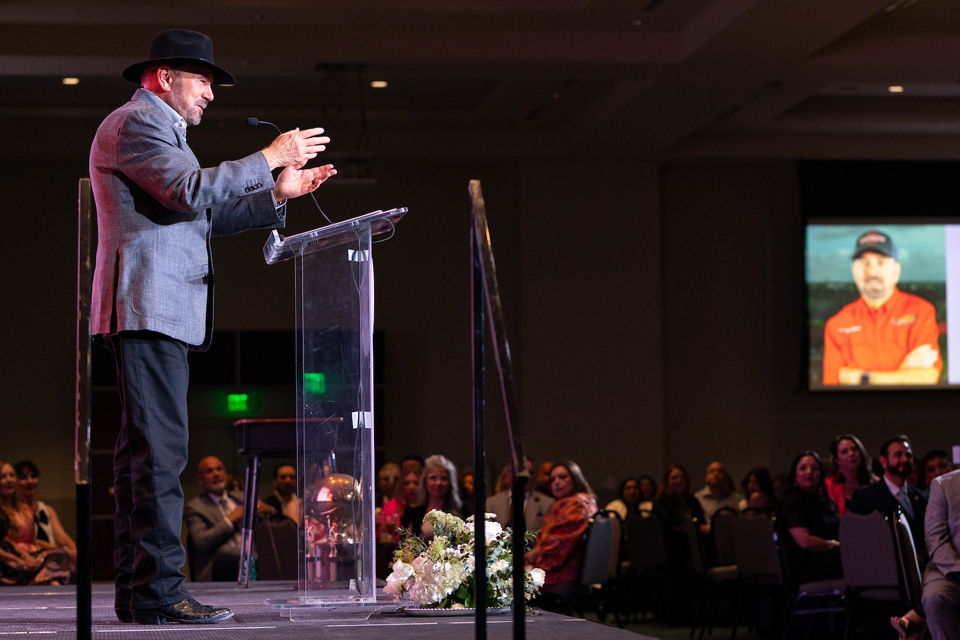 A person wearing a hat speaks at a clear podium on a stage, with a seated audience visible in the background