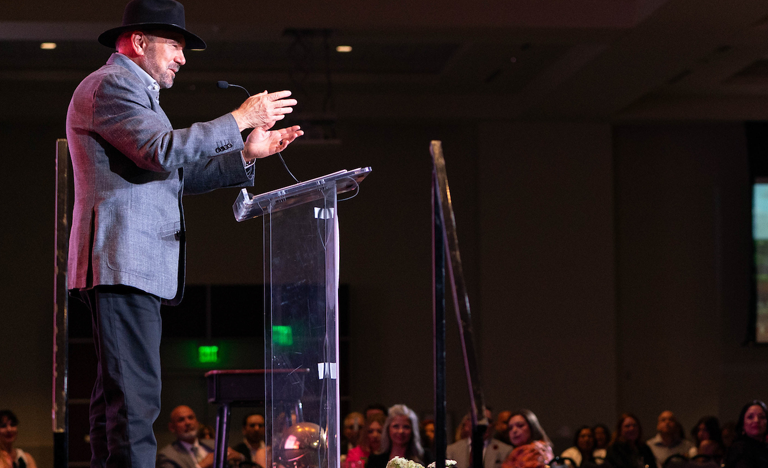 A person wearing a hat speaks at a clear podium on a stage, with a seated audience visible in the background