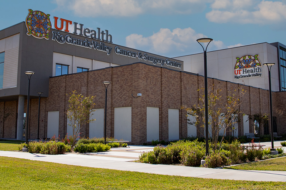 Modern healthcare building labeled "UT Health Rio Grande Valley Cancer and Surgery Center" with large windows, surrounded by green lawns and trees under a blue sky.