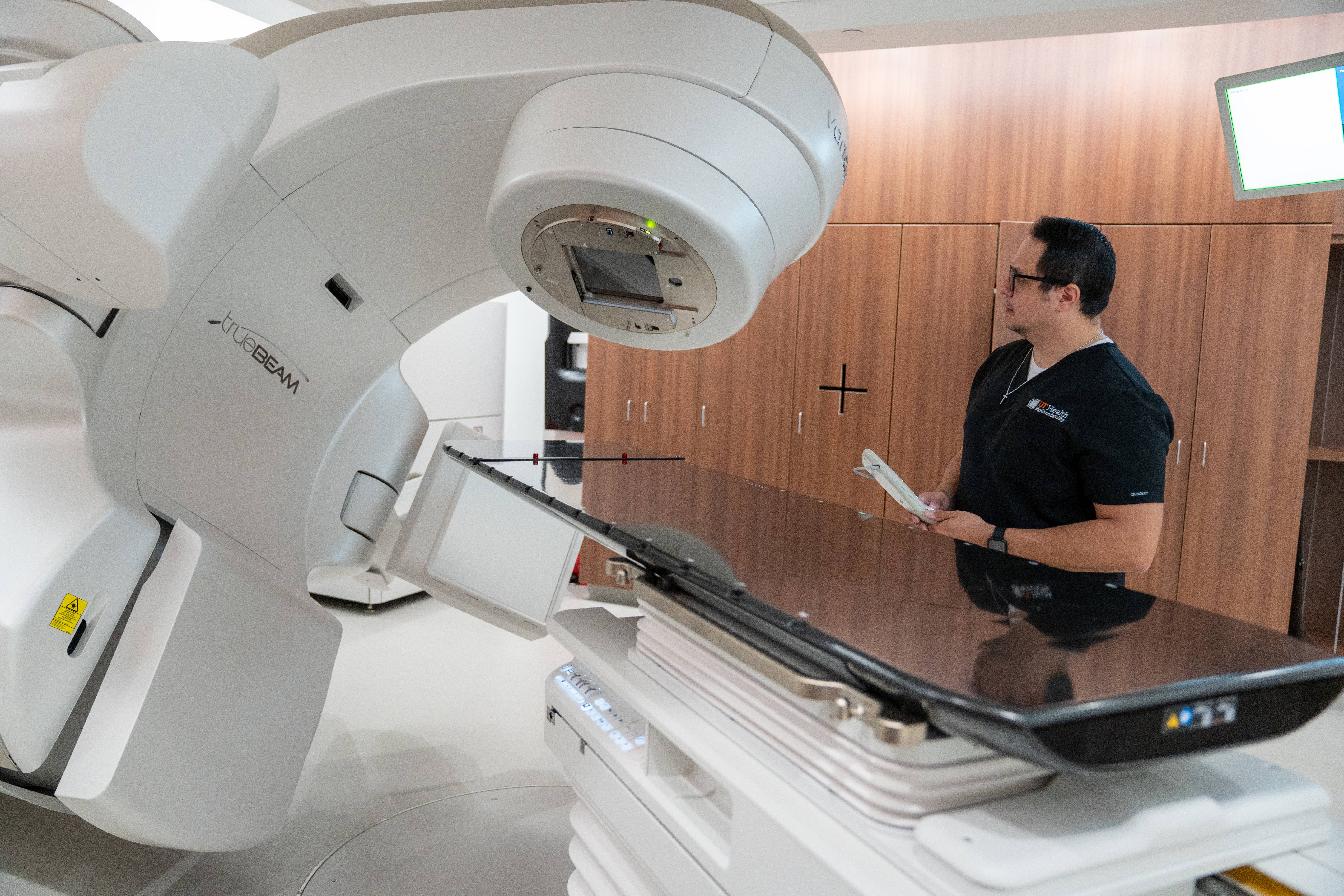 A medical professional in scrubs operates a large, advanced radiotherapy machine in a clinical setting. The atmosphere is focused and high-tech.