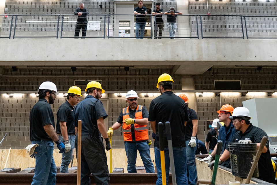 A group of workers in hard hats gather around an indoor rail track while others observe from a balcony above. 