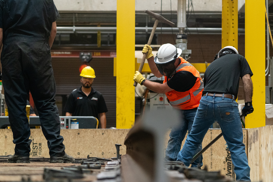 Workers in safety gear install rail components indoors, with one worker using a sledgehammer on the track.