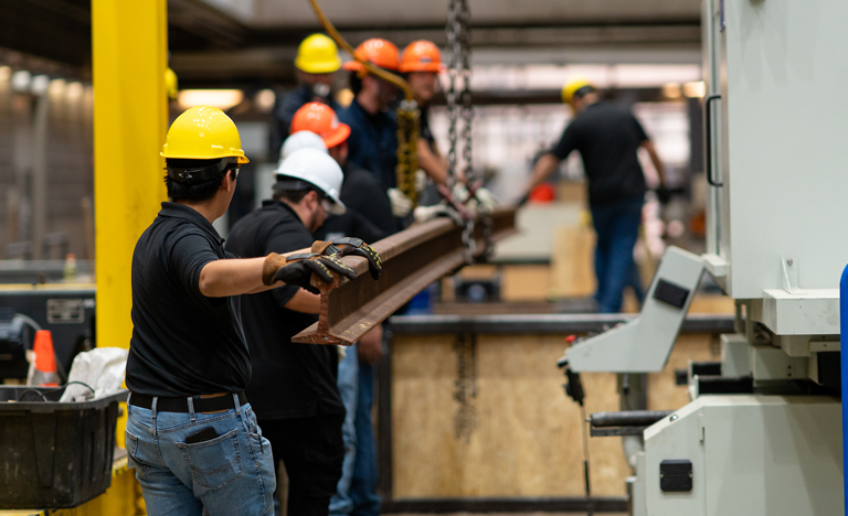 Several workers wearing hard hats guide a long metal rail through industrial machinery inside a factory
