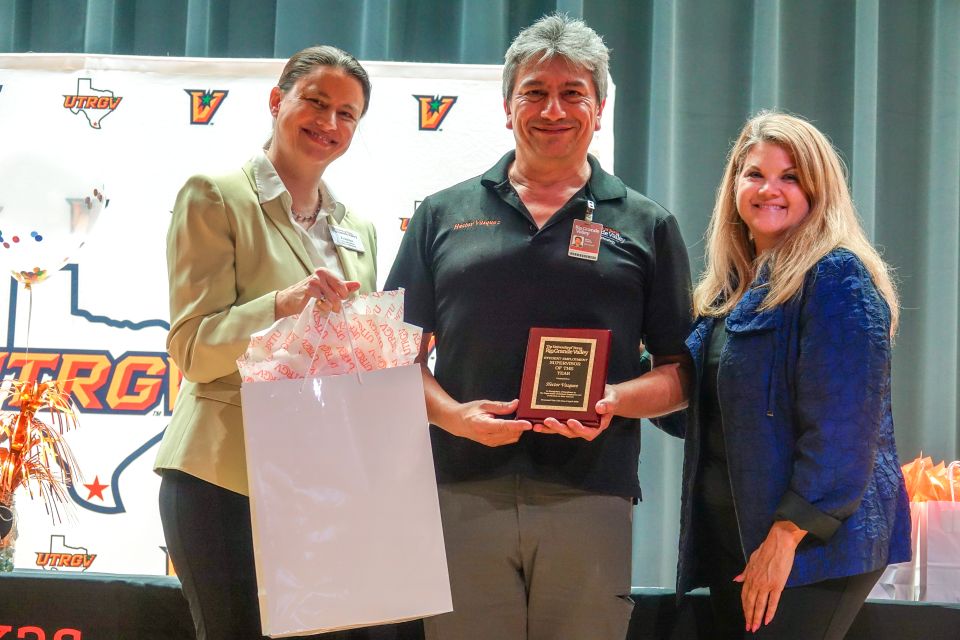 Three persons standing on stage in front of a UTRGV poster behind them, one person accepting an award