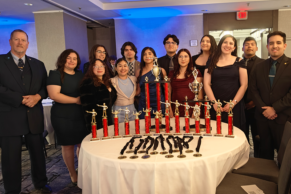 Group of people dressed formally stand behind a round table displaying multiple red-and-gold trophies and medals at an indoor awards event