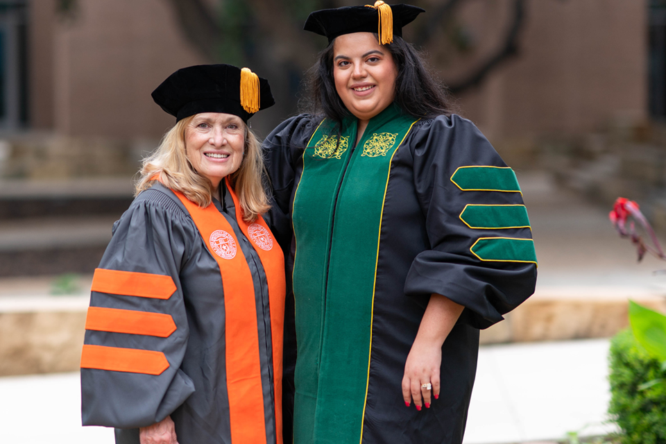 Two people standing close together outdoors on a campus-like setting, both wearing black academic gowns and black tam caps with gold tassels.