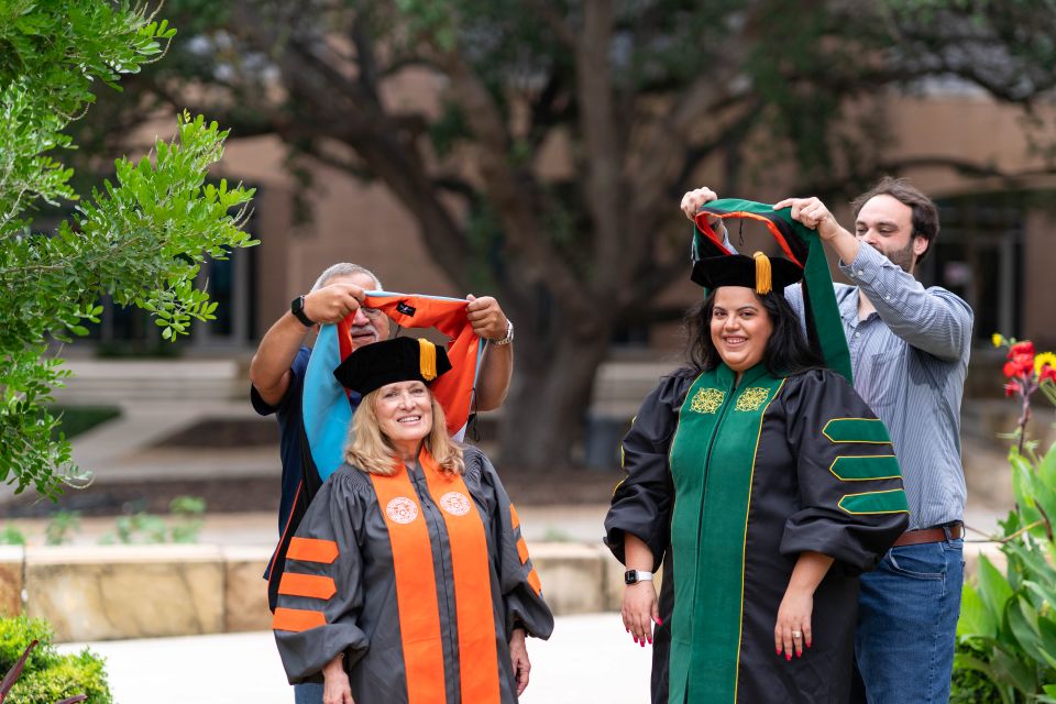 Four people gathered outdoors in a campus setting with trees and a building behind them. Two people in black academic regalia stand in the center