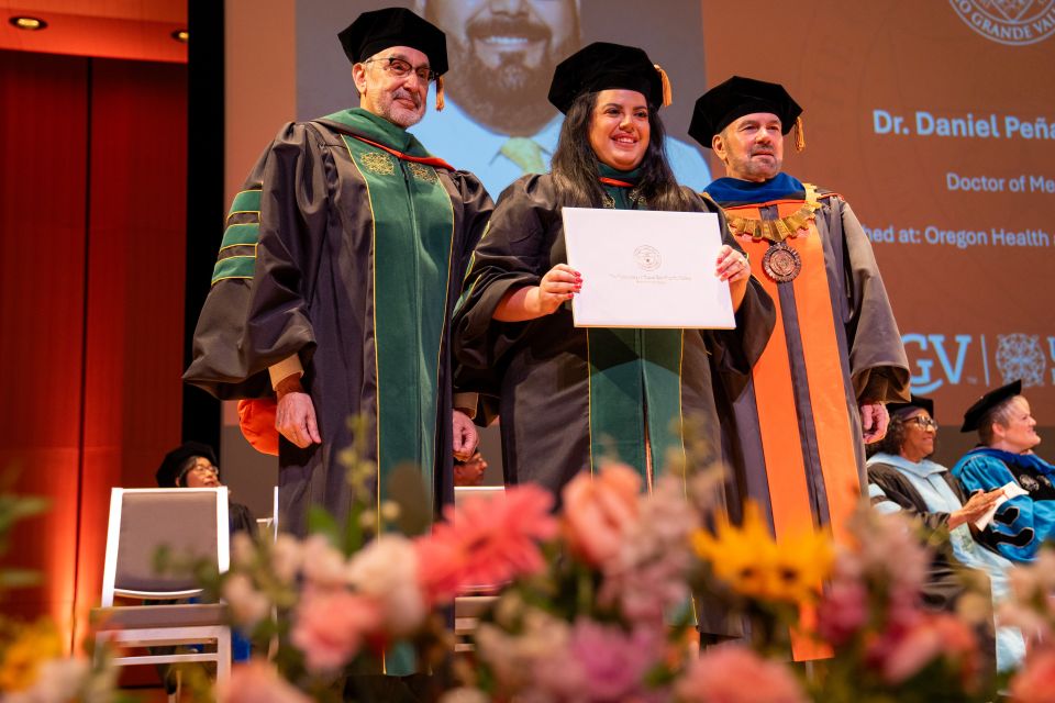 Three individuals in academic regalia stand on a stage during a graduation ceremony. The person in the center, wearing a black gown with green velvet panels, holds a diploma while two others in formal academic gowns stand on either side.