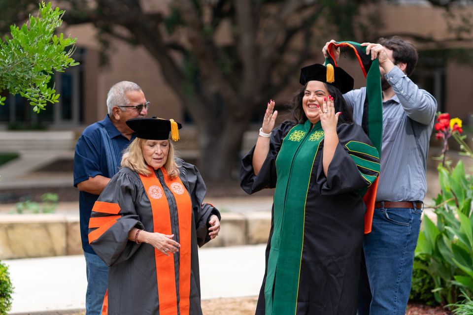Four people gathered outdoors in a campus setting with trees and a building behind them. Two people in black academic regalia stand in the center