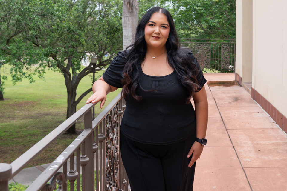  A person with long, dark hair stands outdoors on a walkway beside a building, resting one arm on a decorative metal railing. 