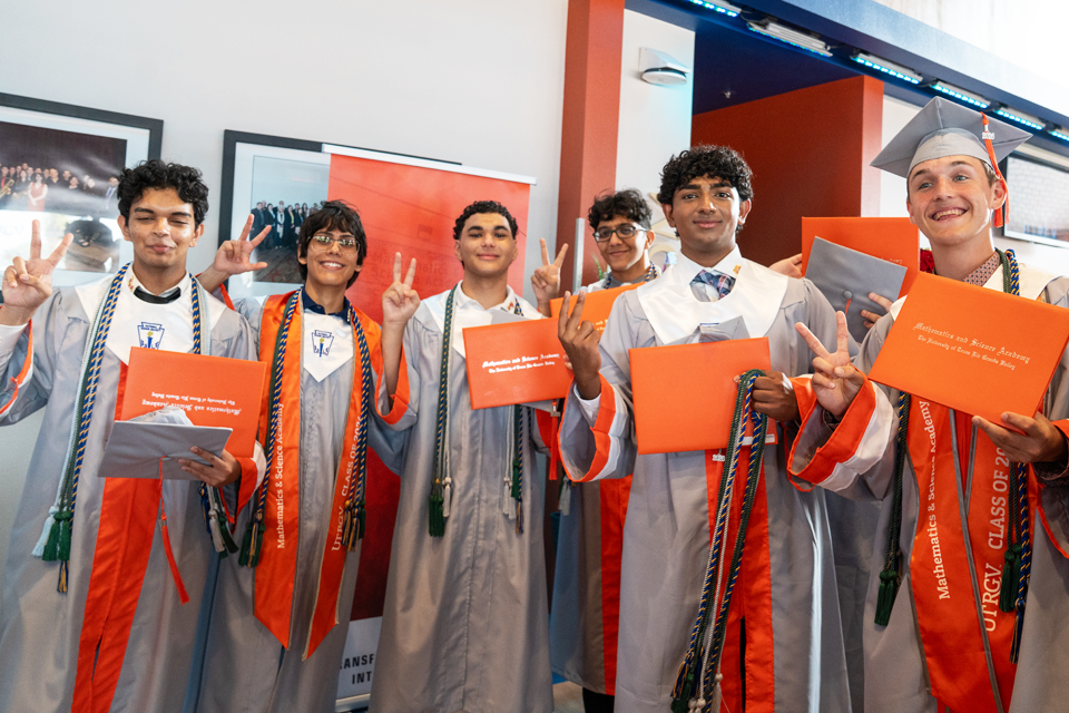 A group of graduates wearing gray caps and gowns with bright orange stoles stand indoors in front of framed photos and a wall with orange accents.