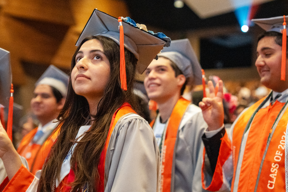 Graduates wearing gray caps and gowns with orange stoles sit closely together at a ceremony.