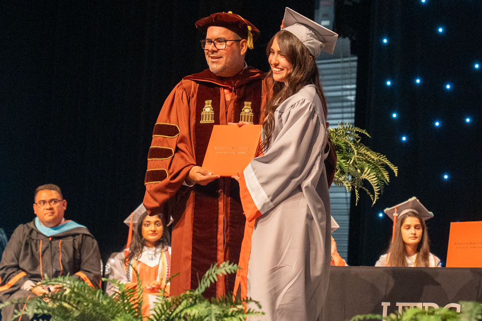 On a stage, a graduate in a gray cap and gown with an orange stole receives an orange diploma cover from a person wearing a maroon academic robe and cap.