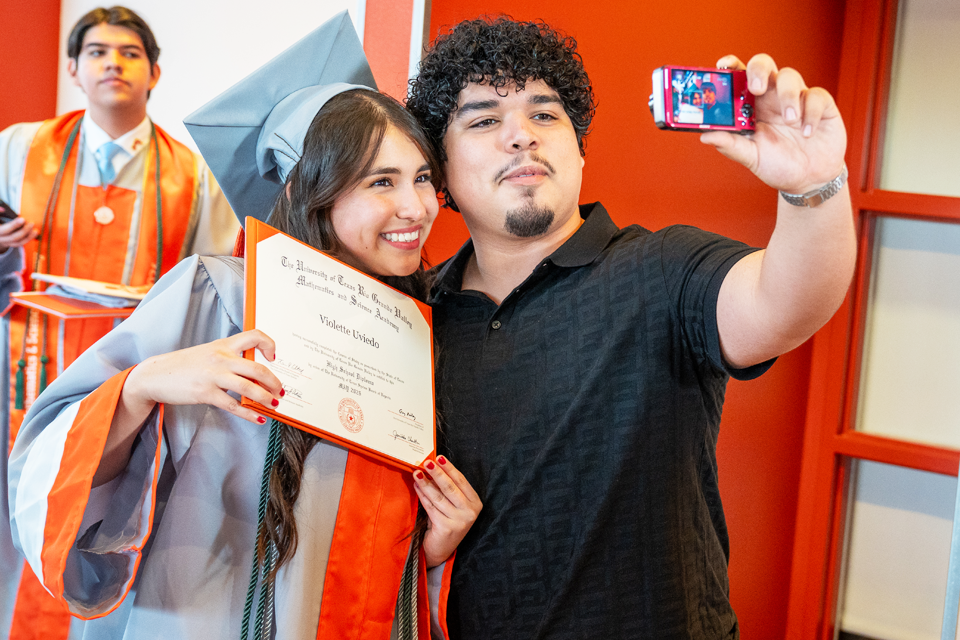 A graduate in a gray cap and gown with an orange stole holds an open diploma while posing for a photo with another person taking a selfie using a small camera.