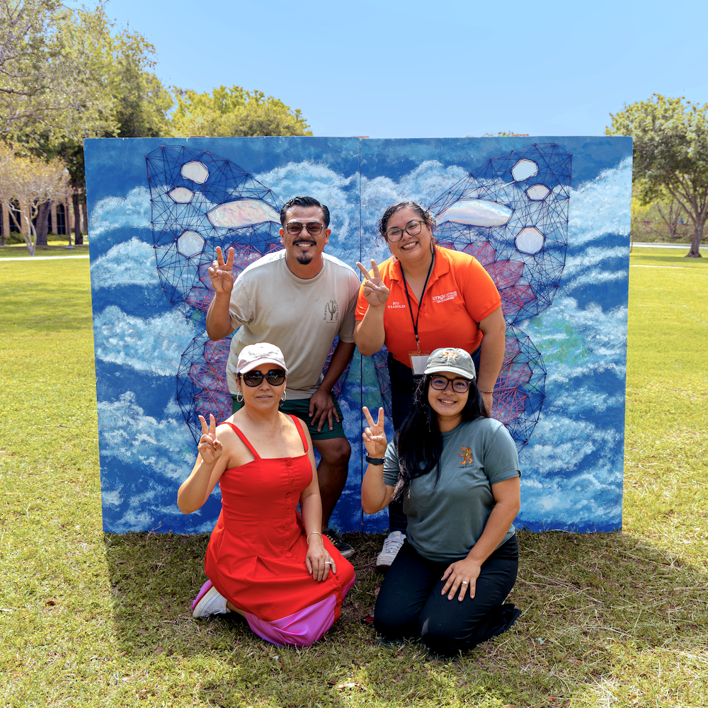Four people pose outdoors on a grassy campus lawn in front of a large painted backdrop depicting a blue sky with abstract butterfly wings made of string art. 