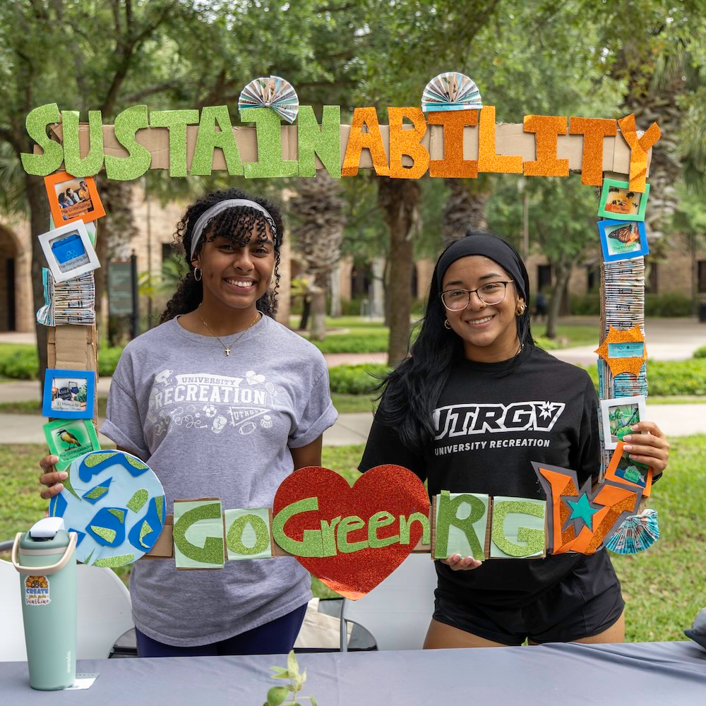 Two people stand behind a table outdoors on a green campus, holding a colorful handmade frame that reads “SUSTAINABILITY” across the top and “Go Green RGV” along the bottom with a red heart in the center.