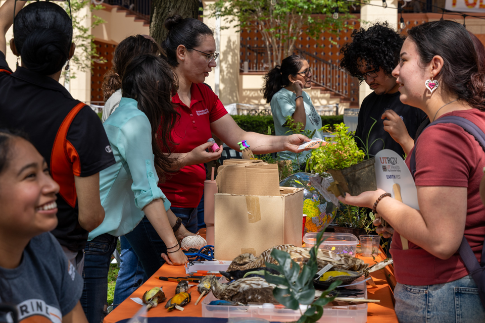 A group of people stands around an outdoor table at a community event, where plants and craft materials are displayed.