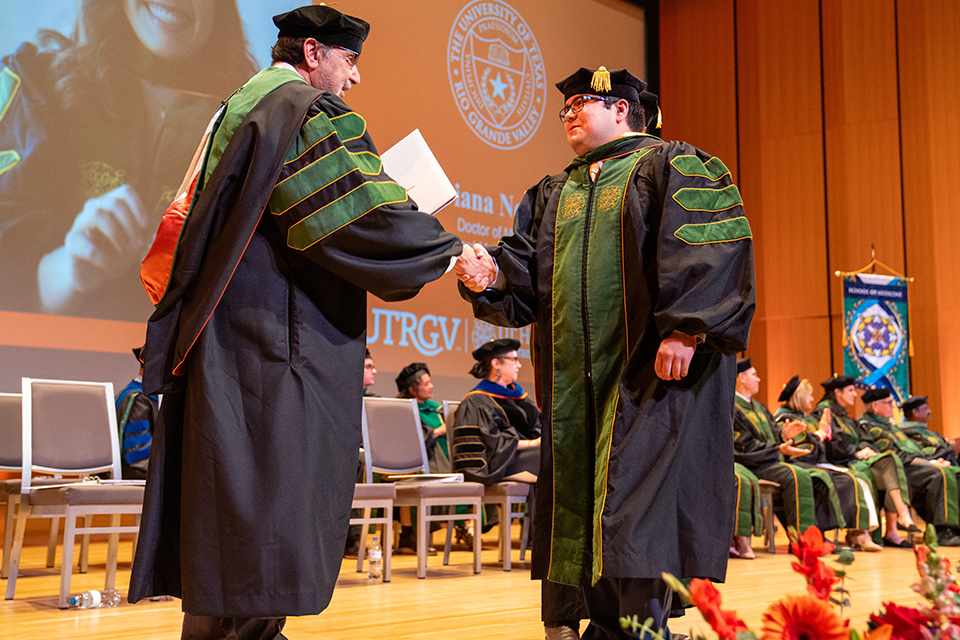 Two people in academic regalia shaking hands on stage. Attendees in similar attire sit in the background. A vibrant, celebratory atmosphere. 