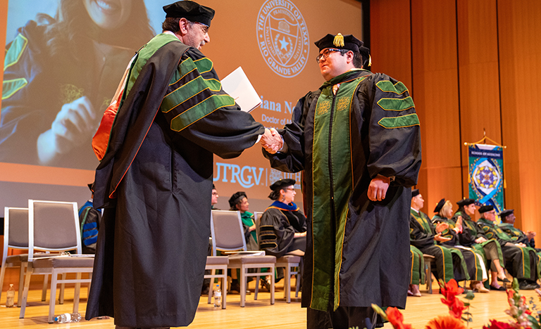 Two people in academic regalia shaking hands on stage. Attendees in similar attire sit in the background. A vibrant, celebratory atmosphere. 