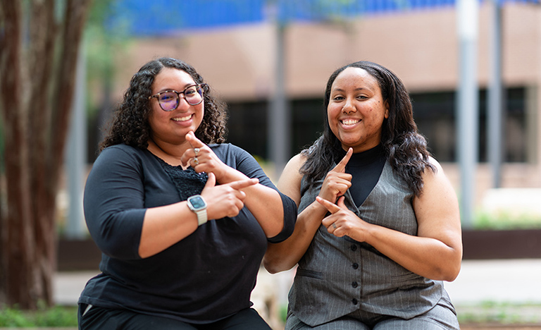 Two women sit together outdoors on the UTRGV campus, smiling and signing in American Sign Language. Gabrielle Black, left, wears glasses and a black outfit, while Alisha Atkinson, right, wears a gray vest and matching pants.