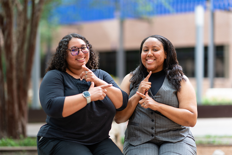 Two women sit together outdoors on the UTRGV campus, smiling and signing in American Sign Language. Gabrielle Black, left, wears glasses and a black outfit, while Alisha Atkinson, right, wears a gray vest and matching pants.
