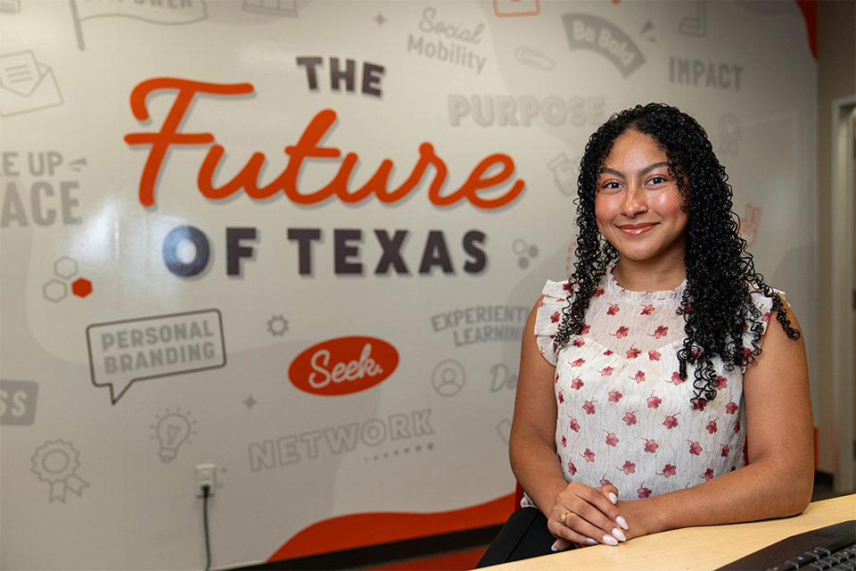Miracle Linnea Sanchez sits smiling at a desk inside the UTRGV Career Center in Edinburg. Behind her is a wall display featuring the words “The Future of Texas” along with career-readiness themed graphics and phrases such as “social mobility,” “network,” and “personal branding.”