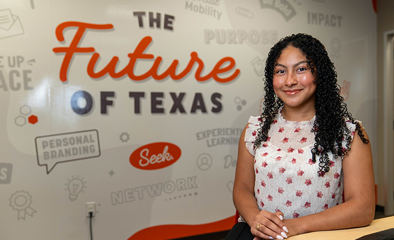 Miracle Linnea Sanchez sits smiling at a desk inside the UTRGV Career Center in Edinburg. Behind her is a wall display featuring the words “The Future of Texas” along with career-readiness themed graphics and phrases such as “social mobility,” “network,” and “personal branding.”