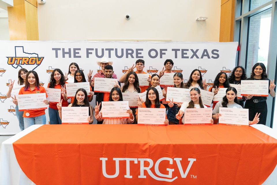 A large group of scholarship recipients pose behind an orange UTRGV table, each holding an oversized scholarship check in front of a backdrop that reads “The Future of Texas.”