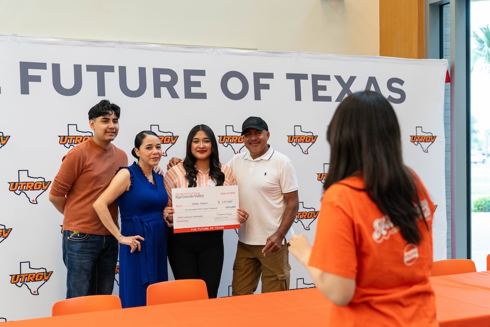 A group of four people stands in front of a “The Future of Texas” UTRGV backdrop, with one person holding an oversized scholarship check while another person in the foreground takes a photo.