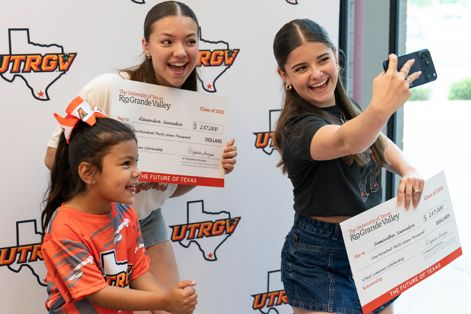 Three people stand in front of a UTRGV step-and-repeat backdrop while holding oversized scholarship checks; one person holds a phone at arm’s length to take a selfie, and another younger child stands in front wearing a UTRGV T-shirt.