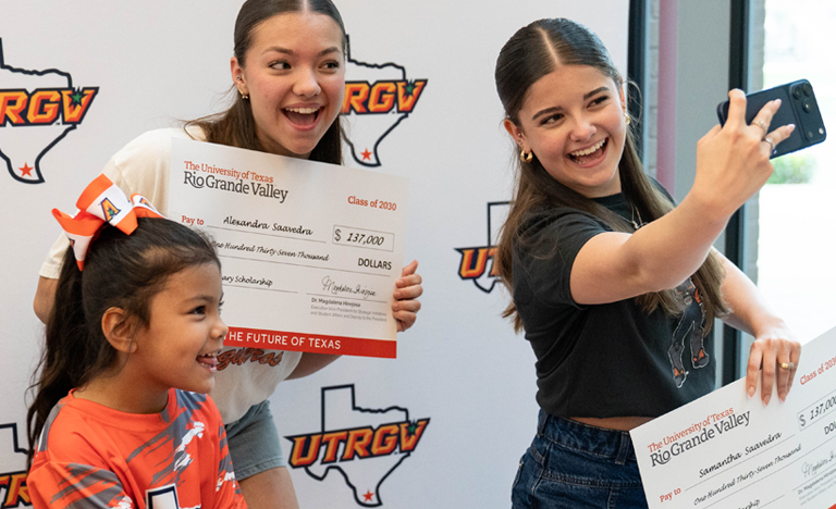 Three people stand in front of a UTRGV step-and-repeat backdrop while holding oversized scholarship checks; one person holds a phone at arm’s length to take a selfie, and another younger child stands in front wearing a UTRGV T-shirt.