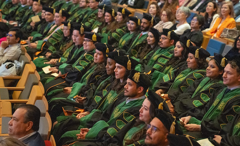 Graduates in black and green gowns seated in a row, ...