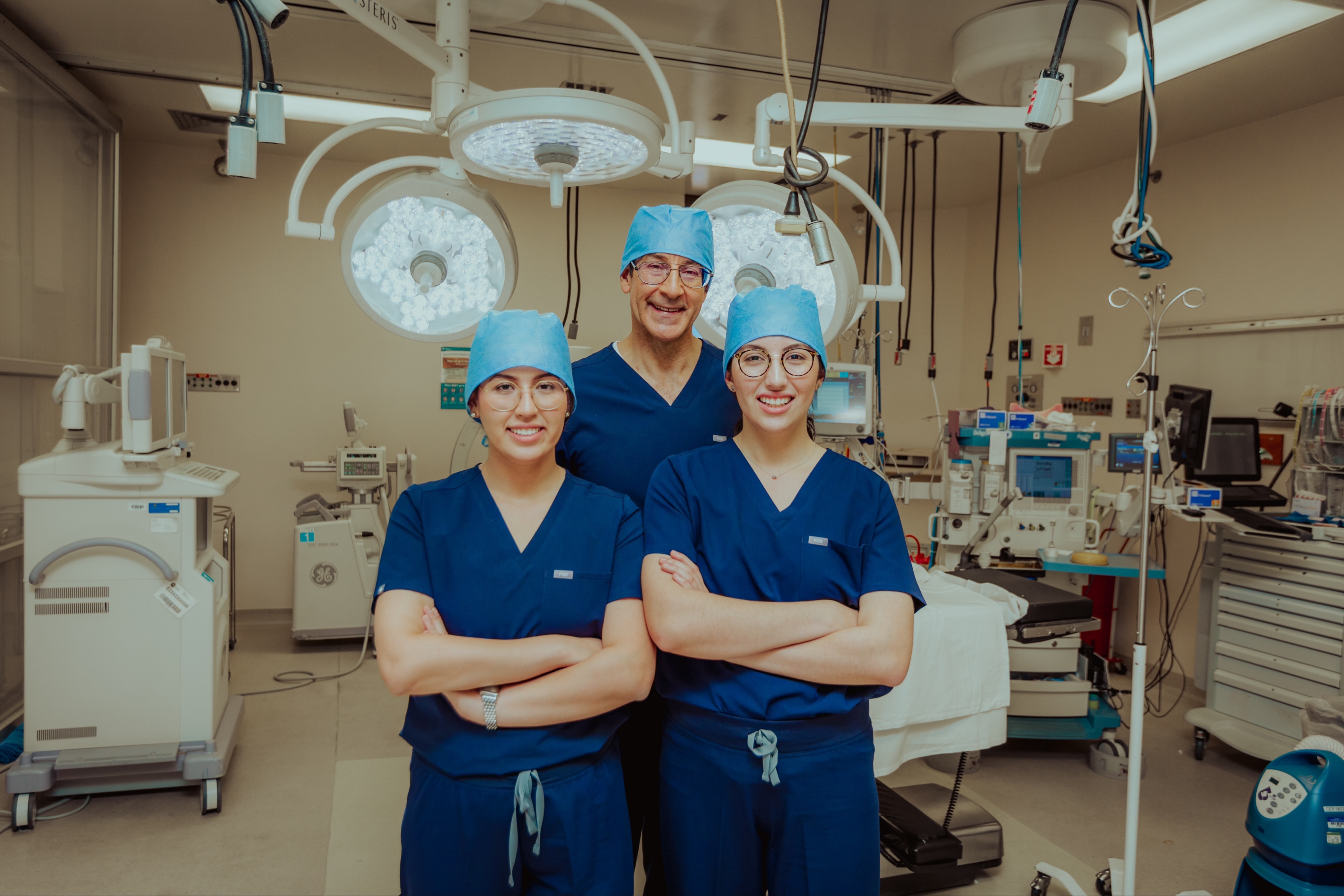 Three medical trainees and a physician in white coats review paperwork together in a hospital or clinical training room.
