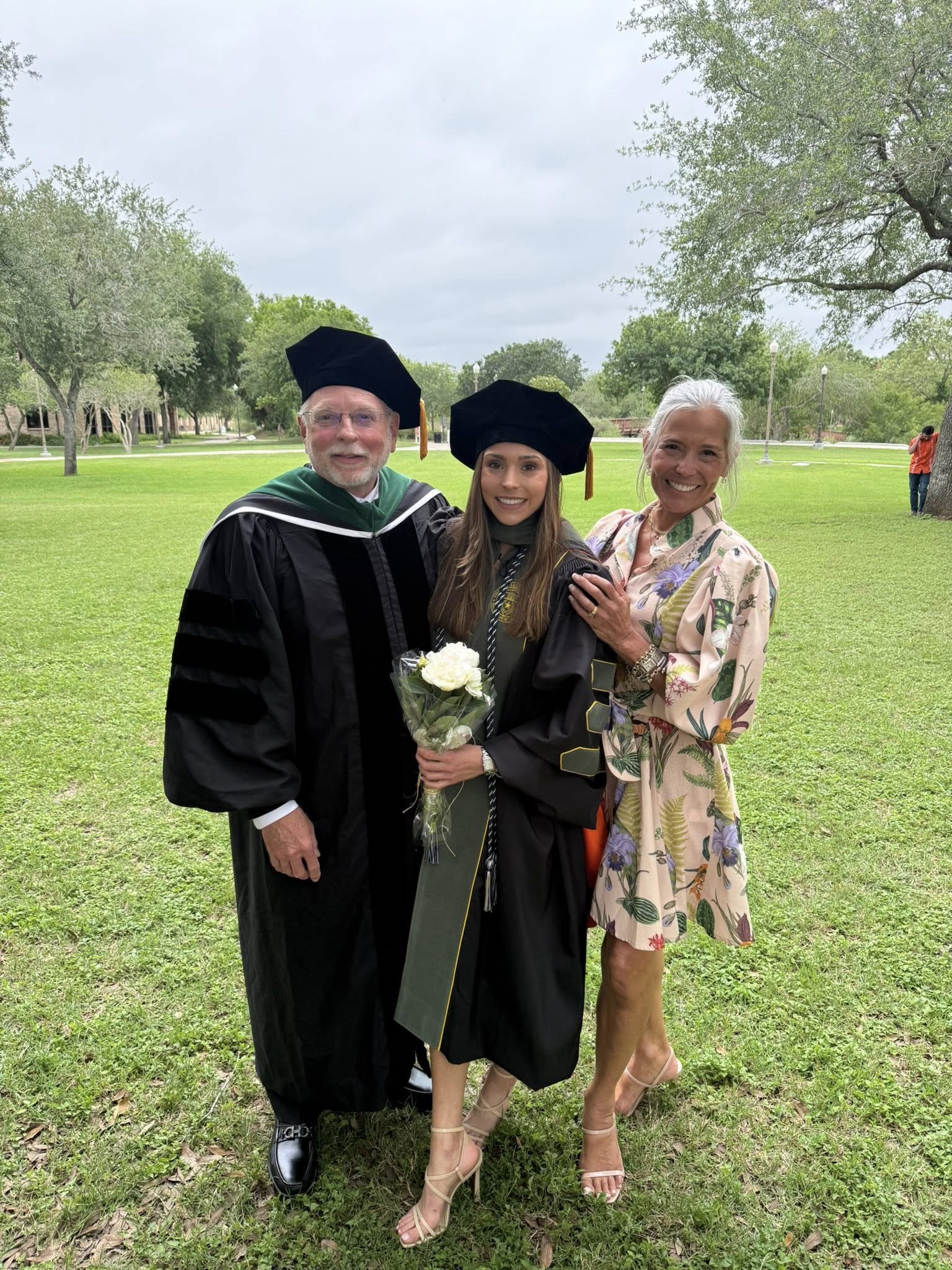 Graduate in cap and gown holding flowers poses outdoors on a grassy campus with two companions.