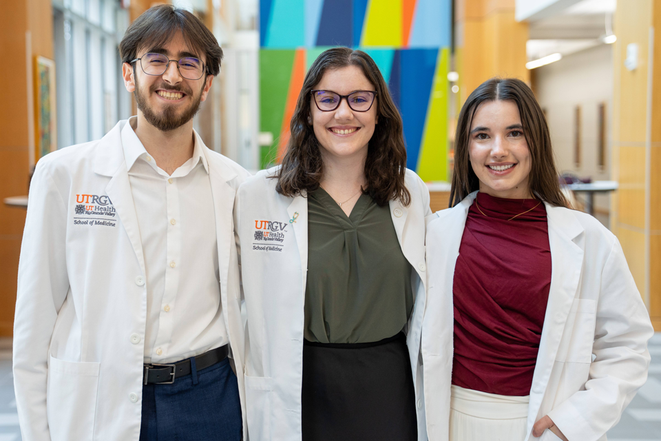 Three young adults in white lab coats stand smiling in a brightly lit hallway with colorful abstract wall art. The atmosphere is welcoming and professional.