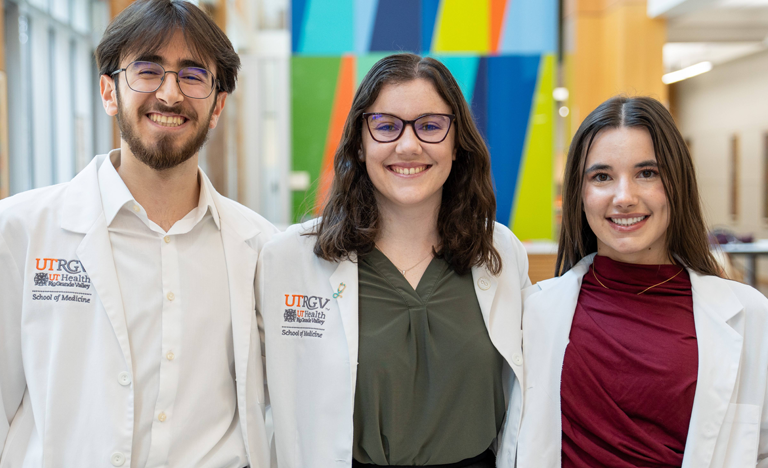 Three young adults in white lab coats stand smiling in a brightly lit hallway with colorful abstract wall art. The atmosphere is welcoming and professional.
