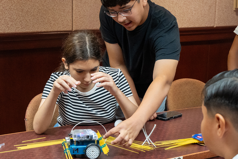 A female student works on putting together a robotic vehicle during a UTRGV summer camp while a male student inspects the vehicle..