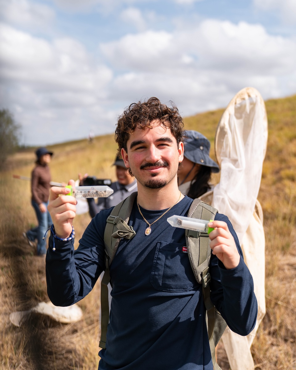 Field research participant holds two specimen tubes containing locusts while standing in a grassy study area.