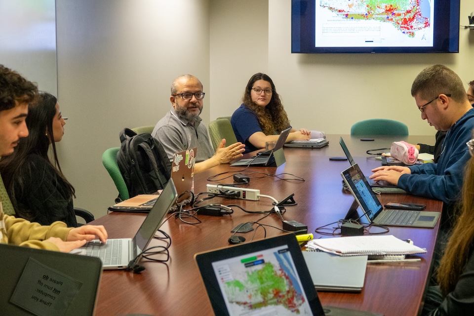 Participants sit around a conference table using laptops while discussing maps and research data displayed on a screen.