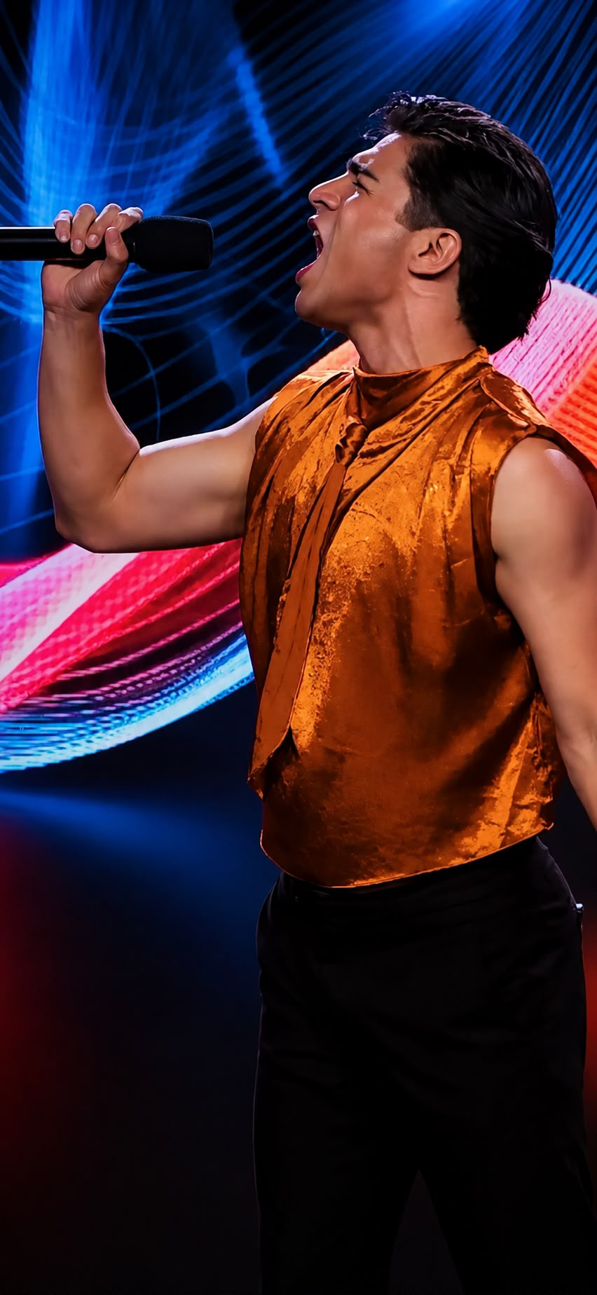 A performer sings into a handheld microphone on stage while wearing a metallic orange sleeveless top. Colorful blue, pink, and purple light patterns fill the background, creating a vibrant concert-style setting