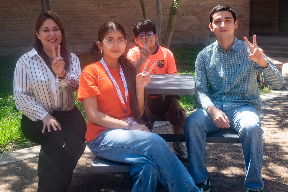 Participants and staff members pose at an outdoor picnic table during a UTRGV camp activity, holding up peace signs in a shaded courtyard.