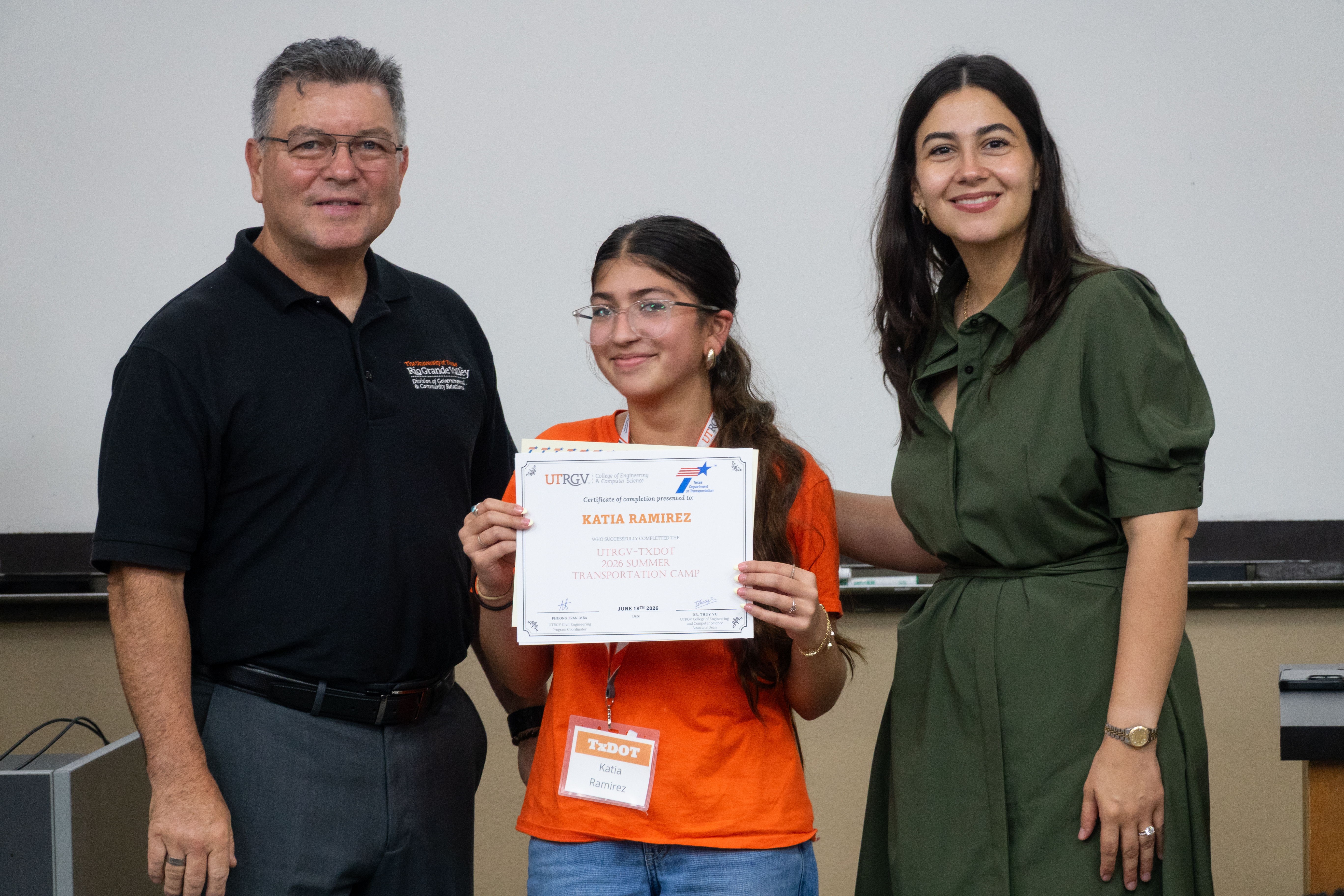 A student in an orange TxDOT camp T-shirt stands between two presenters at the front of a classroom, holding a certificate of completion. The certificate is from the UTRGV College of Engineering and Computer Science and recognizes completion of the 2026 UTRGV–TxDOT Summer Transportation Camp.