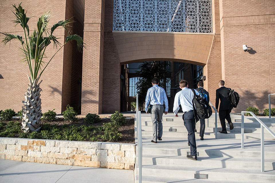 Four people carrying bags walk up a set of concrete steps toward the entrance of a large brick building.