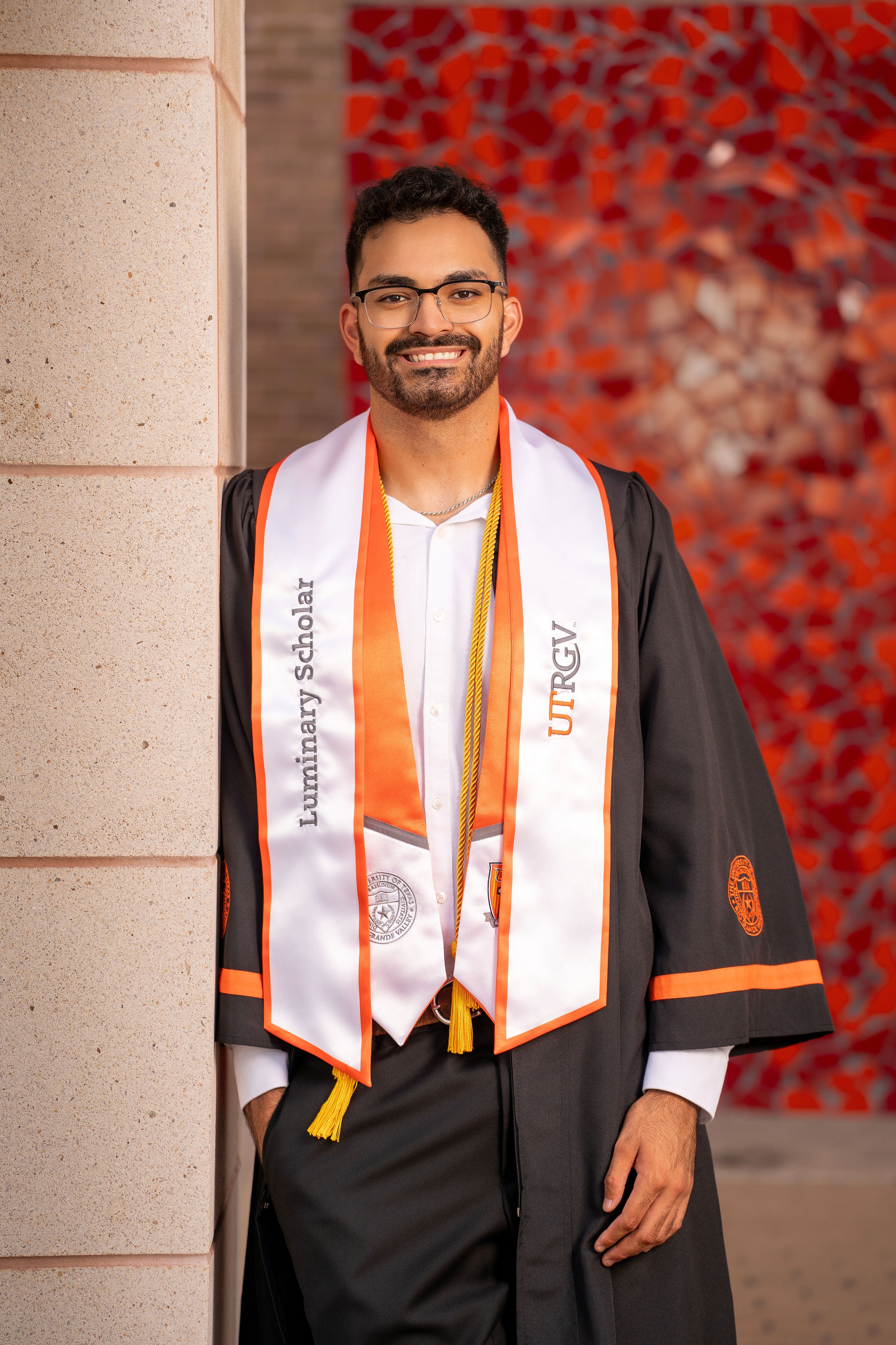 A person wearing a University of Texas Rio Grande Valley graduation gown and a white-and-orange stole standing in front of a brick building